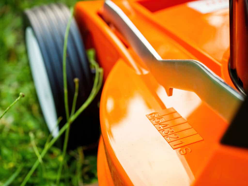 Close-up of an orange lawn mower on grass, highlighting a wheel, metal handle, and stamped serial number 8543215—perfect for maintaining good mowing practices.