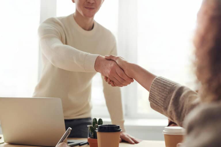 Two people are shaking hands across a desk with a laptop, coffee cups, and a small potted plant on it, suggesting a professional or business meeting.