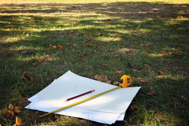 Several sheets of paper with sketches, a yellow tape measure, a pencil, and a ruler lie on grass in a sunlit outdoor setting, with scattered leaves around.